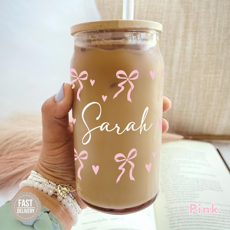 May include: A clear glass reusable iced coffee cup with a bamboo lid and a pink straw. The cup has a pink and white design with bows and hearts and the name "Sarah" written in white. The cup is being held by a hand wearing a white bracelet.