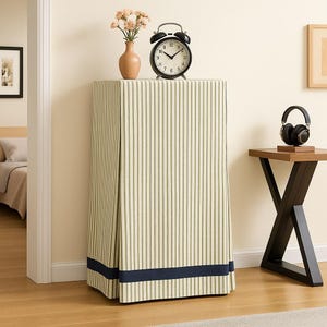 May include: A cabinet covered with a fabric featuring vertical stripes in olive green and cream, trimmed with a navy blue band. A black alarm clock, a vase with flowers, and headphones rest on top. The cabinet is in a room with hardwood floors.