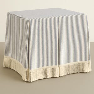 May include: A square ottoman covered in a blue and white striped fabric. The fabric has vertical stripes and a cream-colored fringe along the bottom edge. The ottoman is a neutral piece of furniture.