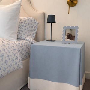 May include: A bedroom scene featuring a bed with floral patterned bedding and a light blue nightstand. The nightstand has a white trim and a small lamp. A framed photo sits on top of the nightstand. A wall sconce with a pleated shade is also visible.