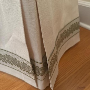 May include: A beige bed skirt with a green leaf pattern border. The skirt has pleats and is made of a soft, textured fabric.