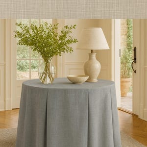 May include: A round, light gray table covered with a matching fabric. A vase of green branches, a small bowl, and a cream-colored lamp with a beige shade sit on top. The table is on a neutral-colored rug.