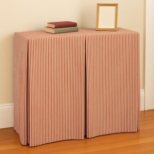May include: A cabinet covered with a red and cream striped fabric. Two books and a gold picture frame sit on top. The cabinet has a pleated front and is placed against a wall and hardwood floor.
