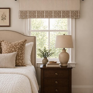 May include: A bedroom scene featuring a bed with decorative pillows and a white quilt. A dark wood nightstand with a lamp, books, and a vase of greenery sits beside the bed. A window is adorned with a striped valance with a geometric border.