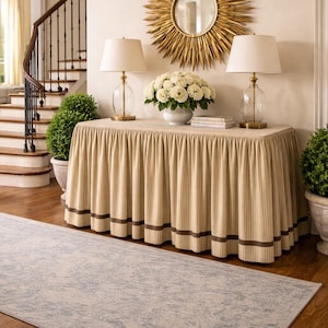 May include: An entryway scene featuring a gold sunburst mirror, two glass lamps with white shades, and a console table with a striped skirt. A vase of white flowers sits on the table, and a light blue rug lies on the wooden floor.