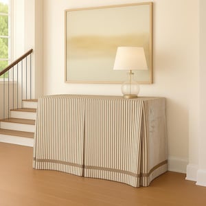 May include: A console table covered with a striped fabric in beige and white. A lamp with a glass base and a white lampshade sits on top. A framed abstract painting hangs on the wall above the table. The room has hardwood floors and a staircase.