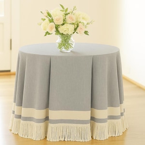 May include: A round, gray tablecloth with a cream-colored band and fringe trim. A clear glass vase holds a bouquet of white and pink flowers. The table is set on a light-colored wood floor.