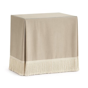 May include: A beige, rectangular table with a fabric cover. The cover has a fringe trim at the bottom. The table is a neutral color, suitable for various interior design styles. The fringe adds a decorative element.