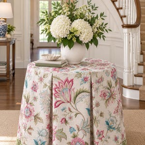 May include: A round table covered with a floor-length, pleated fabric cloth featuring a floral pattern in shades of pink, blue, and green on a beige background, with a green geometric border at the hem and a white vase of white hydrangeas on top.