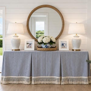 May include: A rectangular console table covered with a blue and white pinstriped fabric skirt featuring a cream-colored fringe trim. The table holds two white ceramic lamps, two white picture frames, and a blue and white bowl of white flowers.