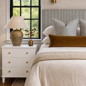 May include: A white three-drawer nightstand with gold knobs, topped with a textured beige lamp and a small gold vase containing dried flowers. Beside it, a bed features a black and white striped headboard, white pillows, and a long velvet pillow.