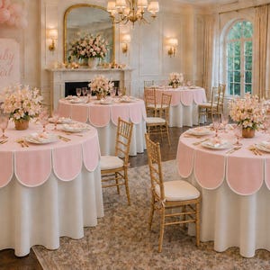 May include: Round tables covered with white floor-length tablecloths and pink scalloped overlays. Each table features floral centerpieces, pink glassware, and white plates. A balloon arch and a sign reading Baby Girl are visible in the room.