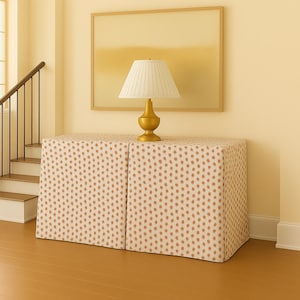 May include: A gold-colored lamp with a white lampshade sits atop a two-piece cabinet. The cabinet is covered in a cream-colored fabric with a repeating pattern of small, orange-brown dots. A painting hangs on the wall behind the lamp.