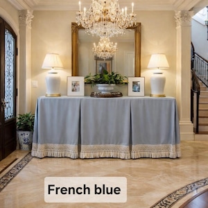 May include: A French blue console table with a pleated design and a cream-colored fringe trim. The table is in a well-lit entryway, with two white lamps and framed pictures on top. The text "French blue" is at the bottom.