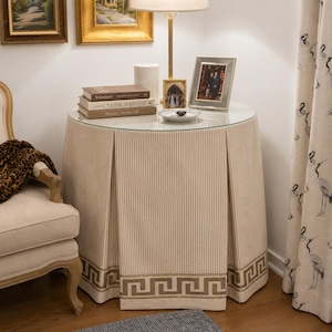 May include: A round accent table with a glass top and a floor-length, pleated beige fabric skirt featuring thin vertical stripes and a dark brown Greek key border pattern along the bottom edge. The table is topped with books and a lamp.