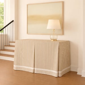 May include: A beige and white striped console table with a white trim. A clear glass lamp with a beige lampshade sits on top of the table. A framed abstract painting hangs on the wall behind the table.