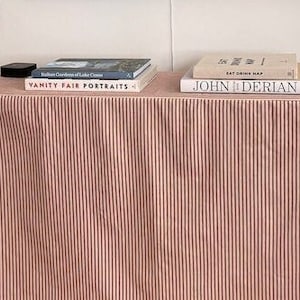 May include: A small table covered with a pink and white striped fabric. The table is topped with books, including "Vanity Fair Portraits" and "John Derian". A small black box sits on the table. The table is against a white wall.