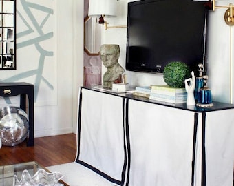 May include: A white media console with black trim, featuring a flat-screen TV, a decorative sculpture, and books. The console is in a living room with a white rug and a glass coffee table.