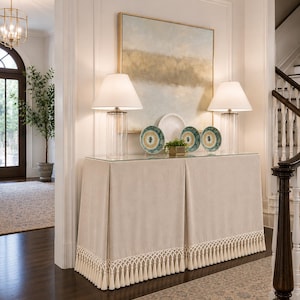 May include: A rectangular console table with a beige fabric skirt featuring a decorative fringe trim with tassels. The table has a glass top, two clear glass lamps with white shades, three patterned plates, and a small green plant in a gold pot.