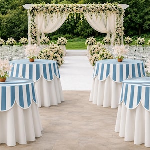 May include: Round tables draped in white floor-length cloths with blue and white striped scalloped overlays. Each table features a central potted arrangement of light pink lilies. The setting includes a white wedding arch and rows of chairs.