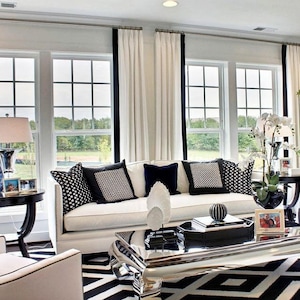 May include: A modern living room with a white sofa, black and white patterned pillows, and a black and white geometric rug. The room features large windows with white curtains and black trim, and decorative side tables.
