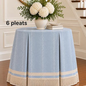 May include: A light blue round table cover featuring 6 pleats, a subtle white dotted pattern, a horizontal beige band near the bottom, and a thick beige fringe trim along the base. The fabric is draped over a circular table surface.