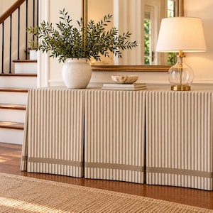 May include: A rectangular console table covered with a floor-length, beige and white striped fabric skirt featuring a solid beige band at the bottom. A white ceramic vase with green foliage, a small bowl, and a table lamp sit on top.