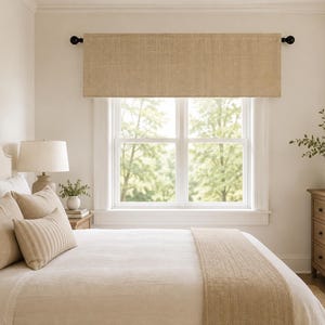May include: A rectangular window valance made of a textured, tan-colored fabric. The valance is mounted on a black curtain rod with round finials, positioned horizontally across the top of a window frame in a bedroom setting.
