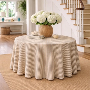 May include: A round table covered with a beige and white striped tablecloth. A woven basket vase holds white flowers. The table sits on a woven rug in a well-lit room with a staircase in the background.