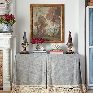 May include: A gray linen tablecloth with a fringe trim, a framed landscape painting, and two decorative black and gold obelisks on a table with a stack of books. The books include titles like "The Great Gatsby" and "Everything I Know About Home".