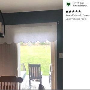 May include: White scalloped valance window treatment hanging over a window with a view of a backyard. The valance is made of a lightweight fabric with a subtle texture.