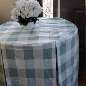 May include: A round table with a white and teal checkered tablecloth. The tablecloth has a pleated drop and is draped over the table. A white vase with white flowers sits on the table.
