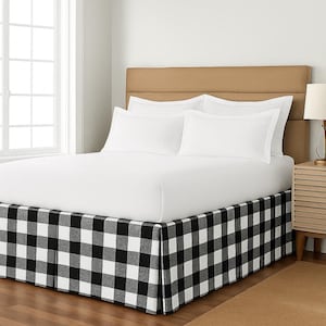 May include: A bed with a tan headboard, white bedding, and a black and white checkered bed skirt. The room has a window and a nightstand with a lamp.