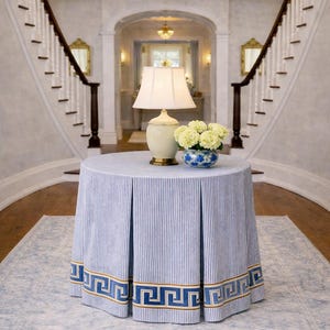 May include: A round table covered with a blue and white striped fabric, trimmed with a Greek key design. A cream-colored lamp and a blue and white floral vase sit on top. The table is in a grand entryway.