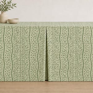 May include: A sage green tablecloth with a repeating pattern of vertical lines and small, irregular dots. The tablecloth is draped over a rectangular table. A vase with greenery and a small bowl are on top.