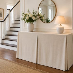 May include: A rectangular console table covered with a floor-length, beige, pleated fabric skirt. On top sits a white ceramic vase filled with white flowers and a white table lamp with a matching ceramic base and a tapered fabric lampshade.