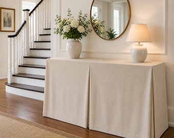 May include: A rectangular console table covered with a floor-length, beige, pleated fabric skirt. On top sits a white ceramic vase filled with white flowers and a white table lamp with a matching ceramic base and a tapered fabric lampshade.