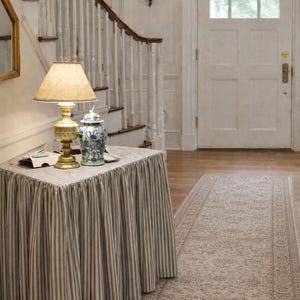 May include: An entryway scene featuring a table with a striped skirt, a lamp, and a blue and white decorative jar. A staircase with a wooden banister and a patterned rug are also visible. The door is white.