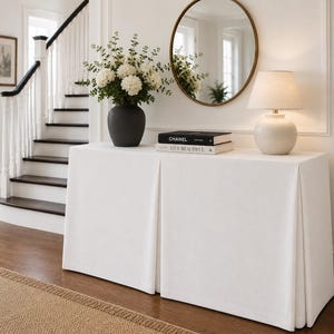 May include: A rectangular console table draped in a white floor-length fabric cover with pleats. On top sits a dark gray vase with white flowers, two stacked books titled Chanel and Live Beautiful, and a white table lamp with a fabric shade.