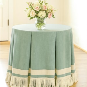 May include: A round table covered with a light teal tablecloth. The tablecloth has a cream-colored band and fringe trim. A glass vase with a bouquet of pink and white flowers sits on top of the table.