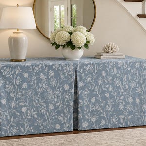 May include: A rectangular console table covered by a floor-length, pleated fabric skirt in a dusty blue color featuring a white botanical floral pattern. The fabric drapes over the table, creating a structured look with two distinct front panels.