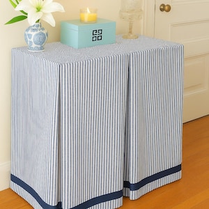 May include: A blue and white striped table skirt with a navy blue border. The table is topped with a light blue box, a vase with a white flower, and a lit candle. A glass cloche sits on the table.