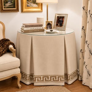 May include: A round accent table featuring a beige fabric skirt with box pleats and a gold Greek key border pattern along the bottom edge. The table has a circular glass top holding three stacked books, a small picture frame, and a lamp.