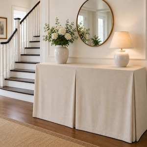 May include: A rectangular console table covered with a floor-length, beige fabric skirt featuring two prominent box pleats. The table surface holds a white ceramic vase filled with white flowers and a white table lamp with a beige fabric shade.