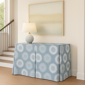 May include: A light blue console table with a white daisy pattern, topped with a light blue lamp with a cream-colored lampshade. A framed abstract painting hangs on the wall above the table. The room has a staircase.