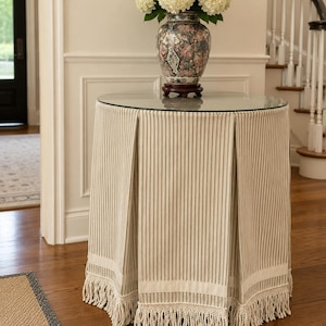 May include: A round accent table covered with a floor-length, beige and white pinstriped fabric cloth featuring box pleats, a horizontal band near the bottom, and a thick fringe trim. A decorative ceramic vase with floral patterns holds white hydrangeas.
