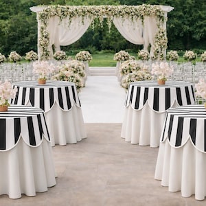 May include: Outdoor wedding reception setup with round tables. The tables have black and white striped tops with scalloped edges and white skirts. Each table holds a small floral arrangement in a brown pot. A floral arch and rows of chairs are in the background.