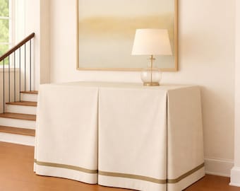 May include: A rectangular console table draped in a floor-length cream fabric cover with a horizontal olive green stripe near the bottom. A glass table lamp with a white drum shade sits on top of the covered table.