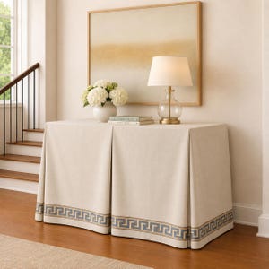 May include: A rectangular console table covered with a floor-length beige fabric cloth featuring a blue and gold Greek key border pattern. On top sits a clear glass table lamp with a white shade, a white floral arrangement, and two stacked books.