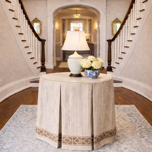 May include: A round accent table with a floor-length, pleated beige and white striped skirt featuring a brown geometric border. On top sits a white ceramic table lamp with a matching shade and a blue and white patterned bowl filled with white flowers.